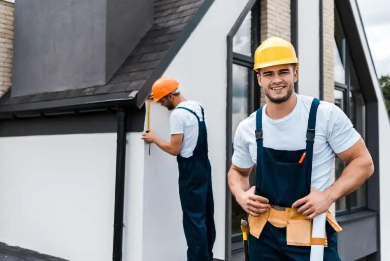 Two men wearing hard hats and overalls stand on the side of a house, likely assessing the construction project.