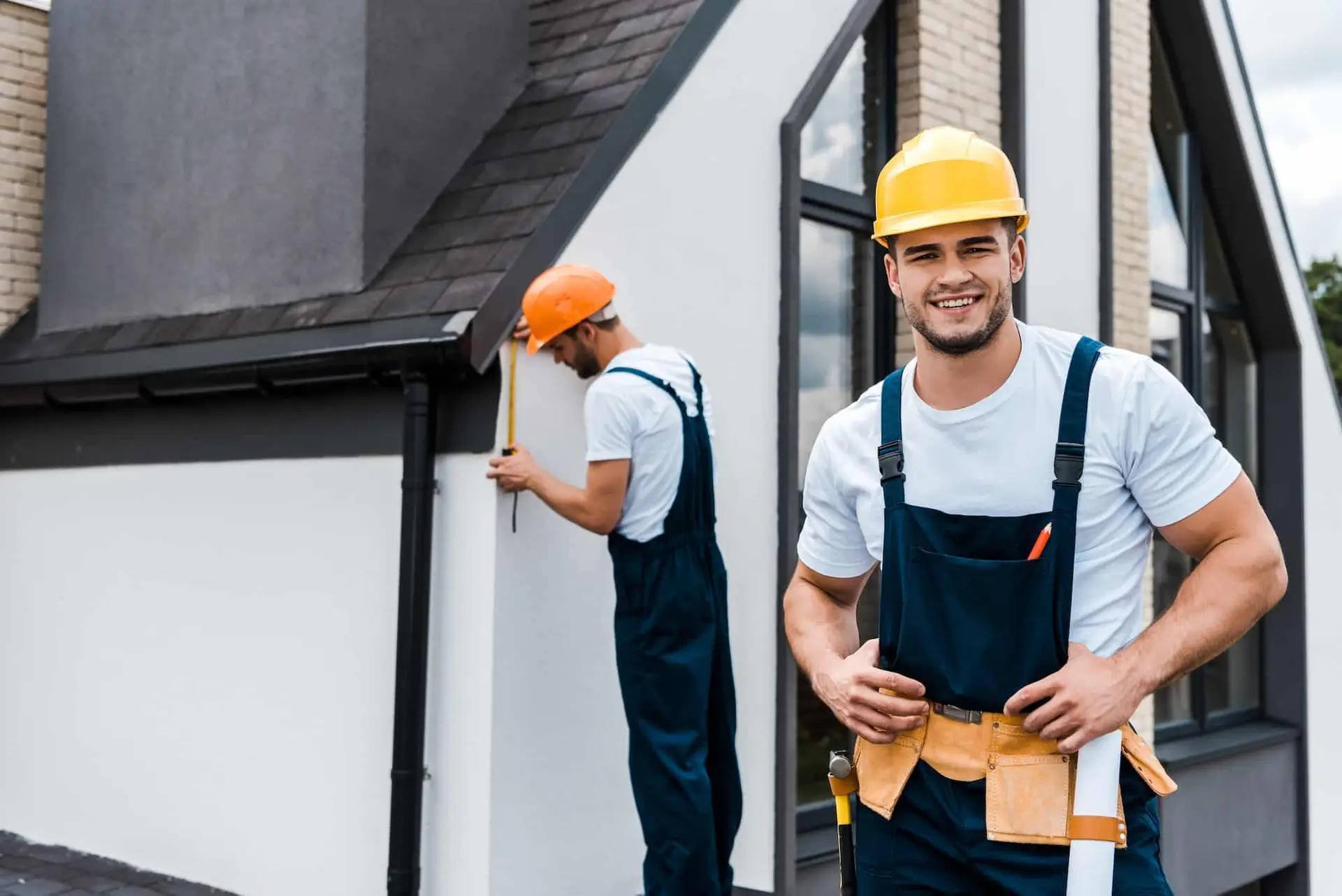 Two men wearing hard hats and overalls stand on the side of a house, likely assessing the construction project.