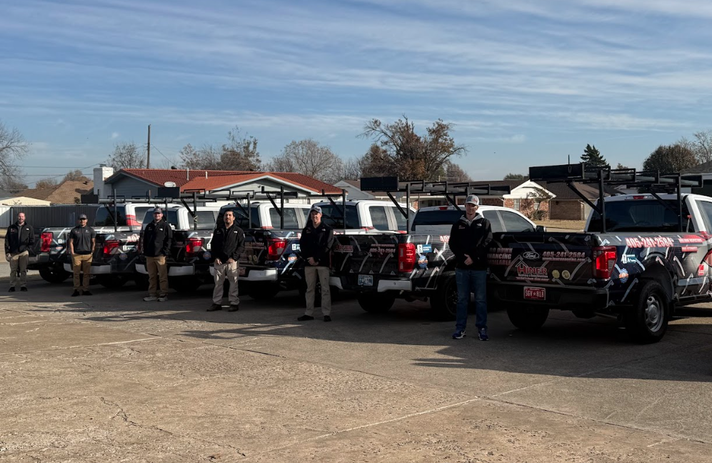Line of black pickup trucks with ladder racks parked in a lot, with several workers standing beside them on a sunny day.
