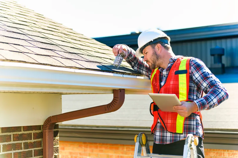 Construction worker in a hard hat and safety vest inspects a roof gutter from a ladder, using a tool and holding a tablet.