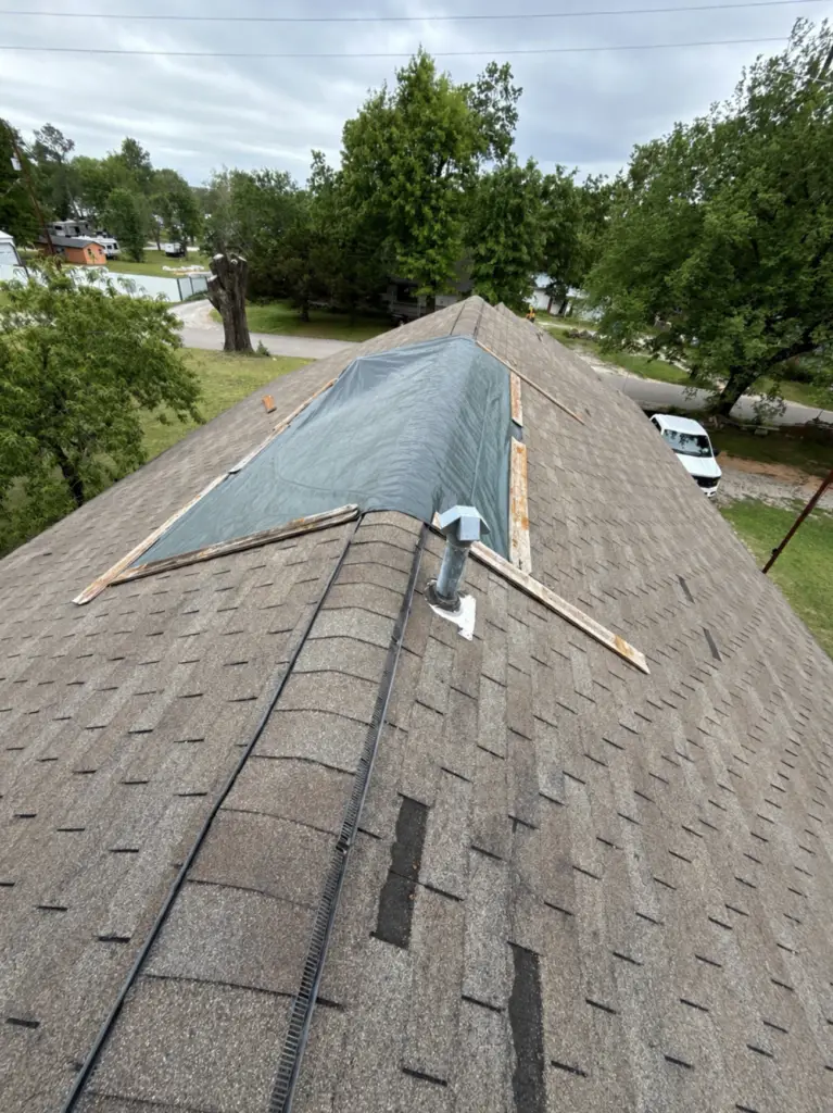 Rooftop under renovation: blue tarp covers a dormer on a brown shingle roof, with wooden planks and a vent pipe visible, trees and houses in the background.