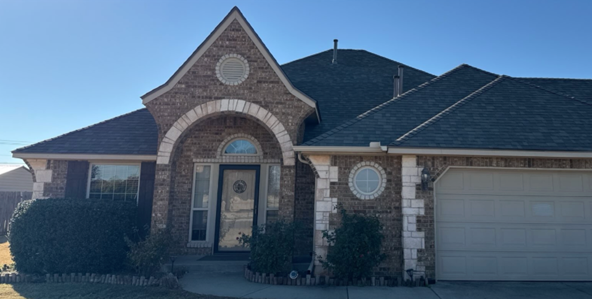 Brick suburban house with arched entryway, circular windows, and a two-car garage with shrubs in front.