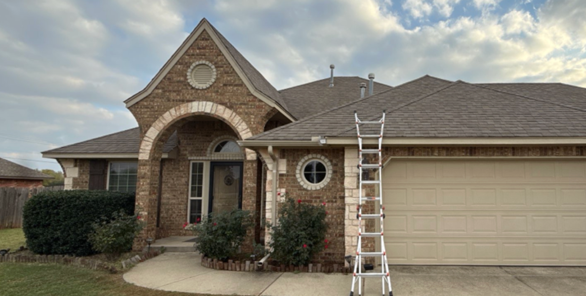 Brick suburban house with arched brick entry, round window, and attached two-car garage; a ladder rests against the roofline, suggesting ongoing maintenance by the front door path framed by bushes and flowers.
