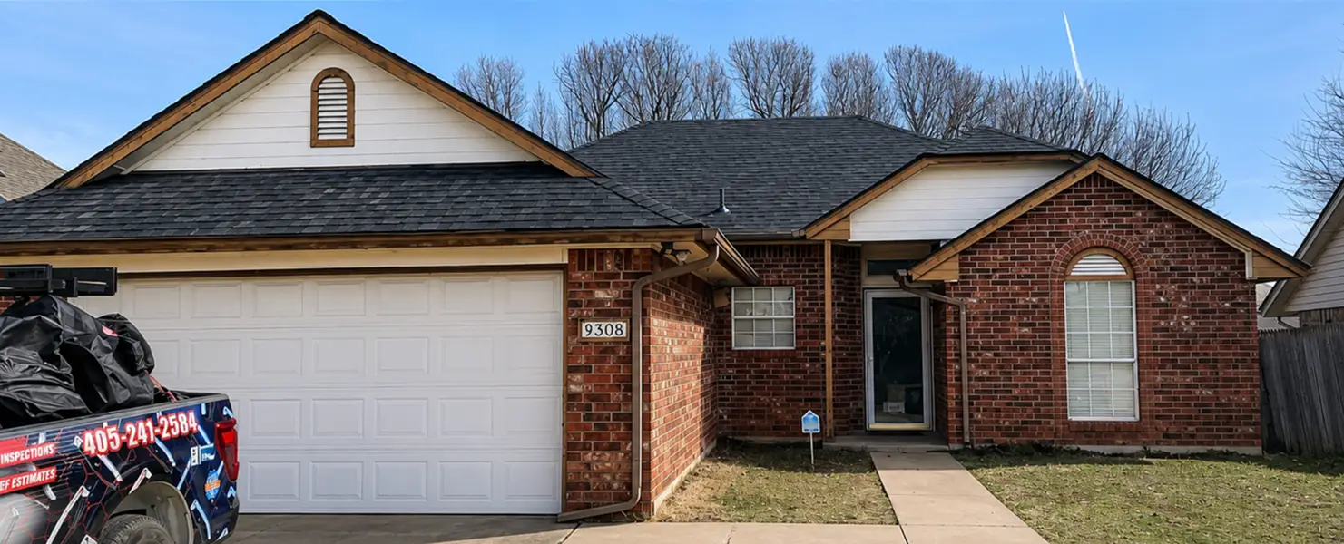 Brick suburban house with a two-car garage and a hemlock-shaded roof; a truck with a tarp is parked at the curb, suggesting a home service visit.