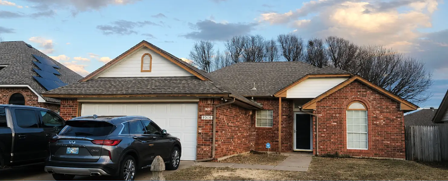 Brick, single-story house with a two-car garage; a gray Infiniti SUV in the driveway and a black van parked beside it under a blue sky.