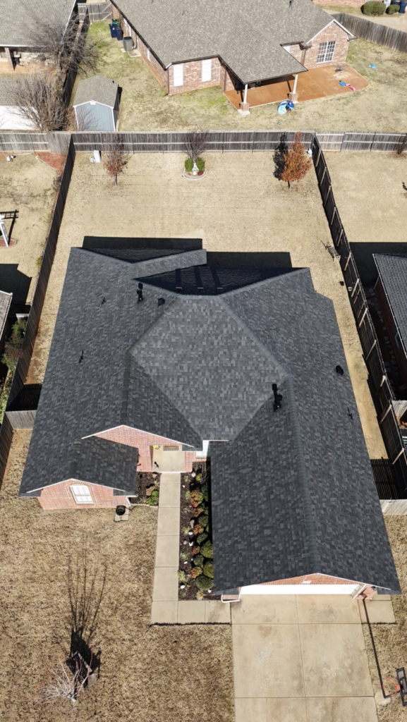 Aerial view of a brick house with a large dark shingle roof, fenced yard, and a concrete driveway nearby.