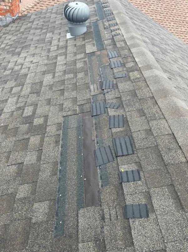 Damaged asphalt shingle roof with a row of small replacement shingles laid along the ridge, near a vent stack; roof shows wear and patchwork along the center line.