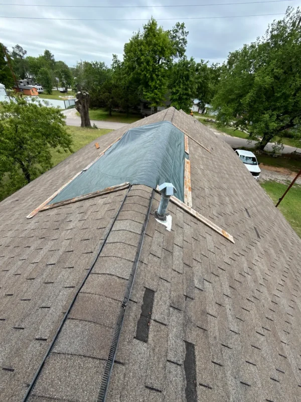 Rooftop under renovation: blue tarp covers a dormer on a brown shingle roof, with wooden planks and a vent pipe visible, trees and houses in the background.